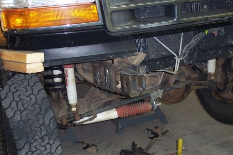 1996 Ford Bronco - Low front-suspension view of a black Ford Bronco raised off the ground, showing the coil spring, white shock absorbers, a red-booted steeri…