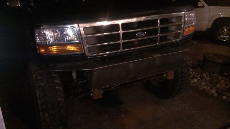 Front of a Ford truck with a chrome grille and aftermarket bumper. Another white SUV is visible to the right