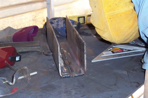 1996 Ford Bronco - A welded steel U-channel or box bracket fabrication sits on a dark workbench, with visible weld beads along its base. Surrounding tools inc…