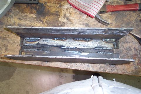 1996 Ford Bronco - A close-up of a welded steel joint showing multiple weld beads joining metal plates, resting on a wooden workbench with a hammer and shop r…