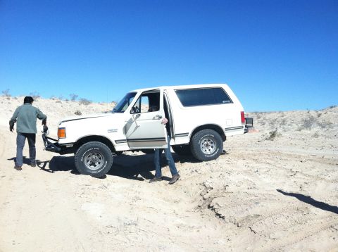 1989 Ford Bronco - A white Ford Bronco is parked in a desert landscape with two people near it.