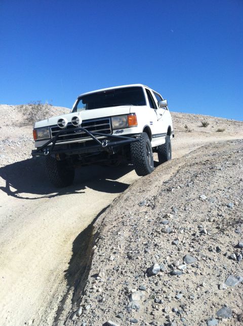 1989 Ford Bronco - White Ford Bronco navigates a dusty, off-road trail under a clear blue sky.