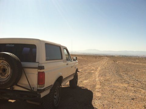 1989 Ford Bronco - A white 1989 Ford Bronco is parked on a dusty, unpaved road with mountains in the distance under a clear blue sky.