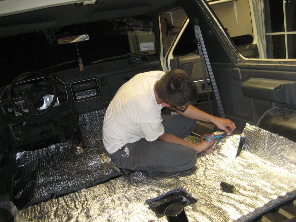 Man cutting silver sound deadening material on the floor of a dark vehicle interior. Steering wheel and dashboard visible