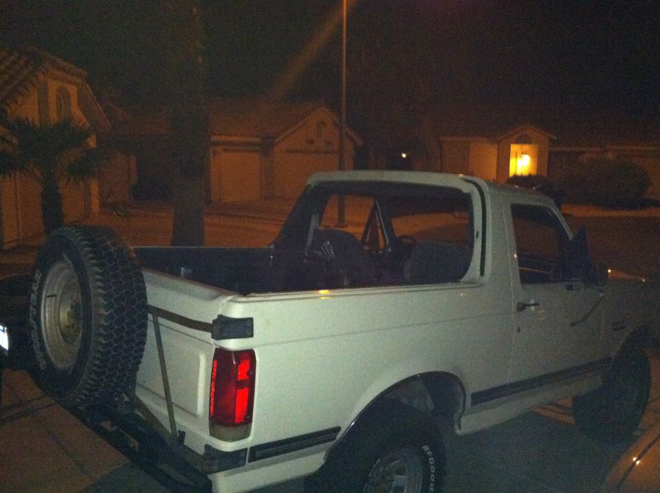 White Ford Bronco, rear view, with spare tire mounted on swing-away carrier. Roll cage visible inside