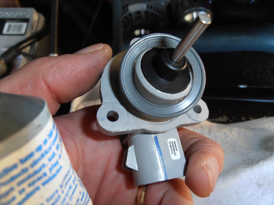Hand holding a gray brake master cylinder component with a metal rod inserted. Label reads 'SENSOR ASSEMBLED IN MEXICO'