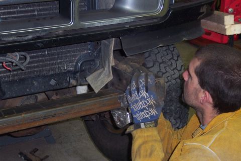 1996 Ford Bronco - A man wearing black 'Mechanix' work gloves and a tan/yellow welding jacket works underneath the front end of a dark vehicle, viewing the fr…