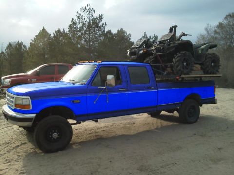 1994 Ford Bronco - A blue Ford F-Series truck is parked on a dirt lot, carrying two ATVs in its bed. Another truck is visible behind it.