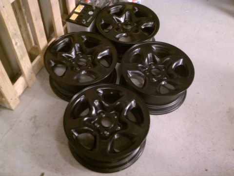 1998 Toyota Camry - Set of four black alloy wheels stacked indoors, with a car battery and wooden pallet visible in the background.
