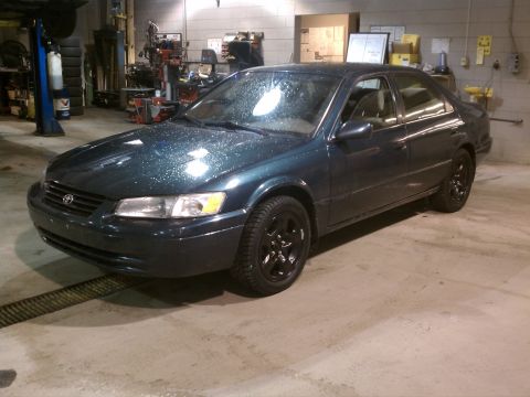 1998 Toyota Camry - A dark green 1998 Toyota Camry with black rims sits in a garage, possibly undergoing maintenance.