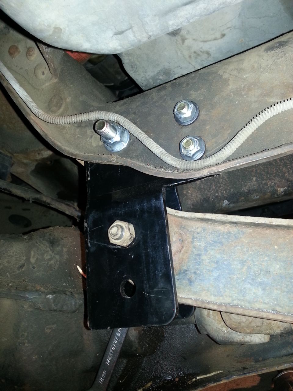 Undercarriage view of a vehicle with a black metal mounting bracket attached to a rusty metal beam, secured by bolts. A wrench is visible