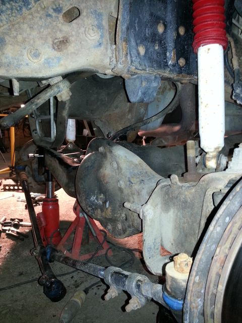 1994 Ford Bronco - The underside of a vehicle is shown, with suspension parts and a jack supporting it. A website logo is visible at the bottom.