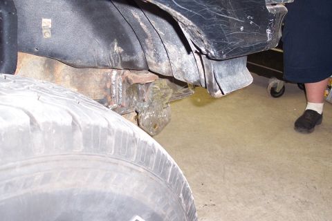 1996 Ford Bronco - Underside view of a vehicle's rear frame and body area showing surface rust on the frame rail, a black plastic fender liner/heat shield, an…
