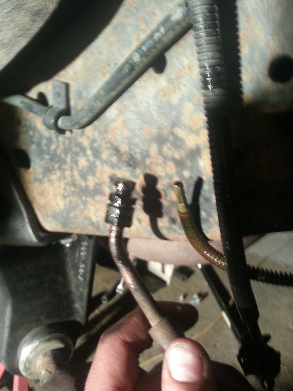 Close-up of rusted vehicle frame with brake lines and a hand holding a metal tube