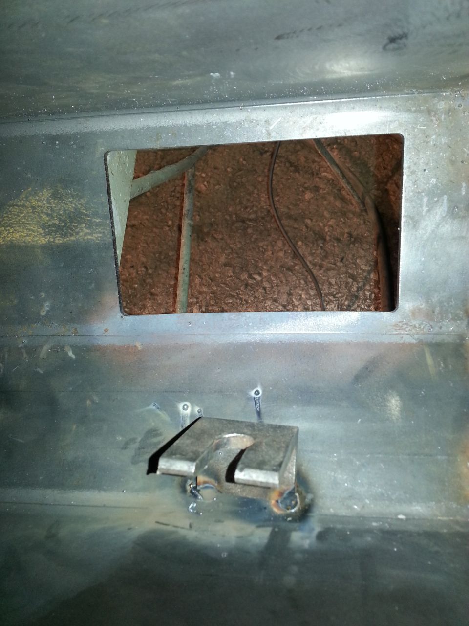 Close-up of a metal bracket welded to a vehicle frame, with a rectangular opening revealing insulation and wires