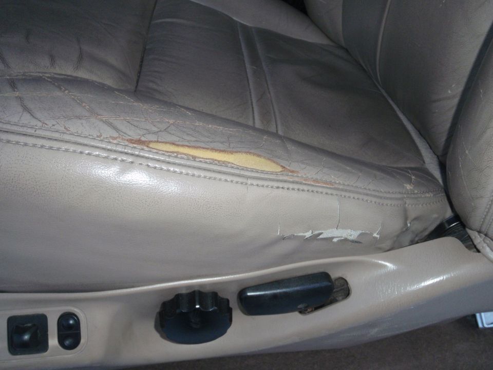 Close-up of worn tan leather car seat with tear on the side cushion and cracked leather. Seat adjustment controls visible