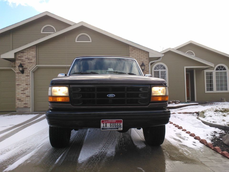 Front view of a dark-colored Ford F-150 truck parked in a driveway with snow on the ground. The grille has a prominent Ford emblem