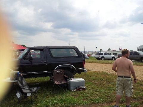 1986 Ford Bronco - A black Ford Bronco is parked on grass with a cooler and chairs nearby. A shirtless man stands in the foreground looking away from the came…