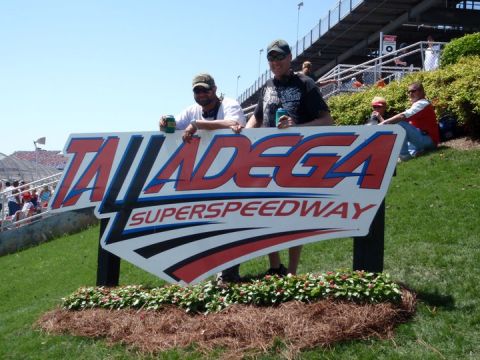1986 Ford Bronco - Two men pose with a large Talladega Superspeedway sign. The sign is white with red and blue lettering and accents. In the background, grand…