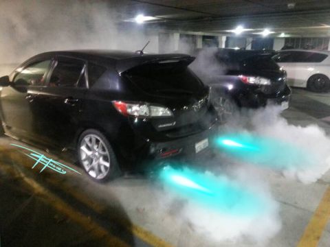 1989 Mazda MX-6 - Two vehicles emit bright blue exhaust flames in a parking garage, creating a smoky scene.