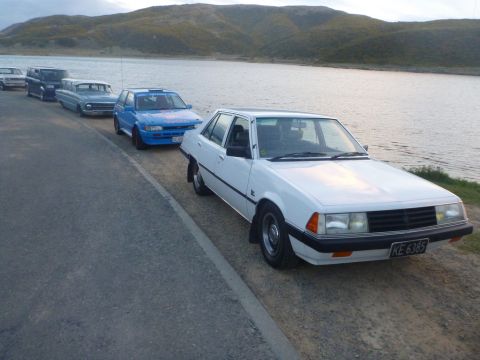 1969 Lincoln Continental - Several cars are parked in a line along the side of a road next to a large body of water with hills in the background.