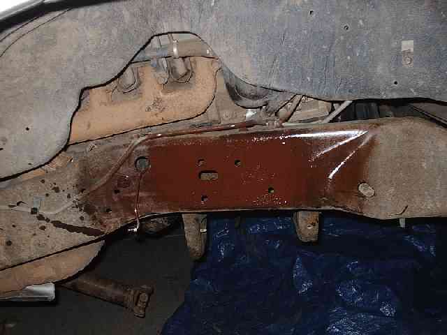 Frame crossmember under a Bronco painted with reddish-brown rust primer, blue tarp below