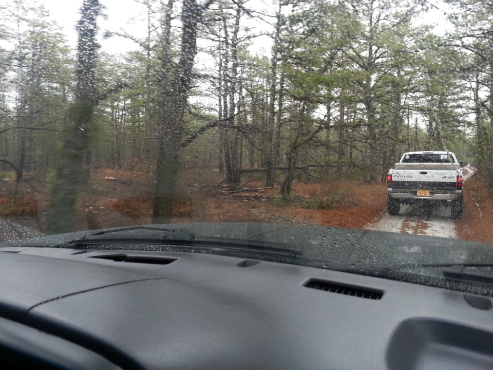 View from inside a vehicle looking at a white Dodge pickup truck driving away on a wet dirt road through a pine forest