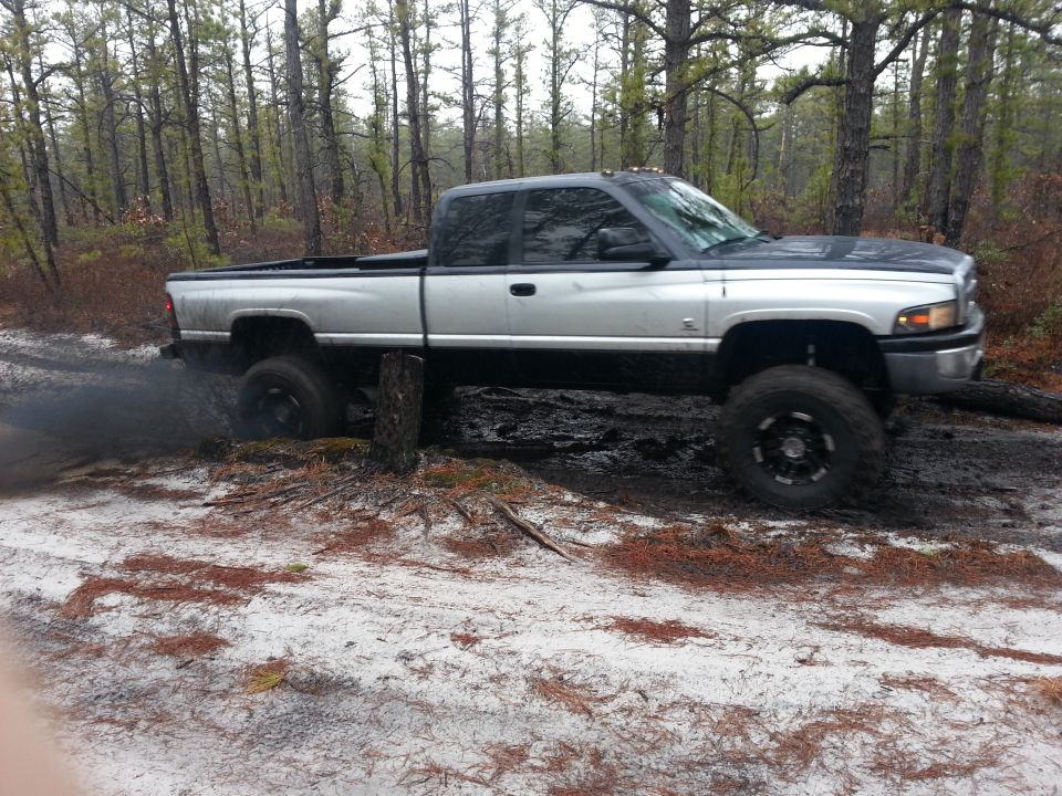 Silver and black Dodge Ram pickup truck stuck in mud and dirt in a wooded area, kicking up black smoke from the rear