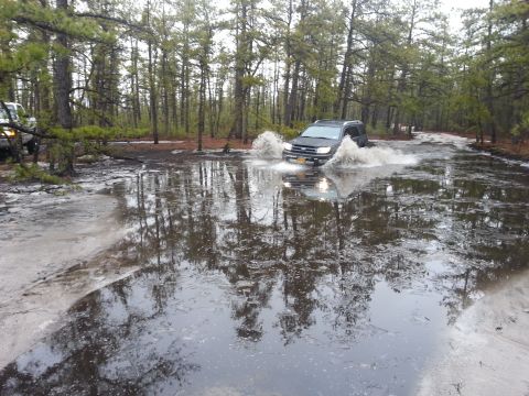 1987 Toyota 4Runner - A dark SUV navigates a flooded path in a wooded area, with another vehicle partially visible on the left.