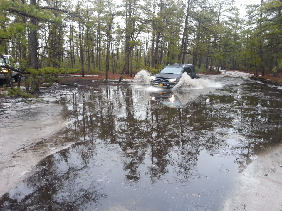 Dark SUV splashing through water on a forest road; another vehicle visible to the left