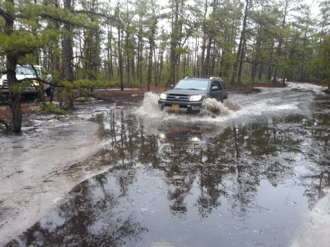 1987 Toyota 4Runner - Two SUVs driving through water in a wooded area, one kicking up a large splash.