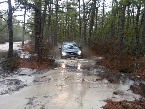1987 Toyota 4Runner - SUV navigates a muddy trail in a wooded area, kicking up water and dirt.
