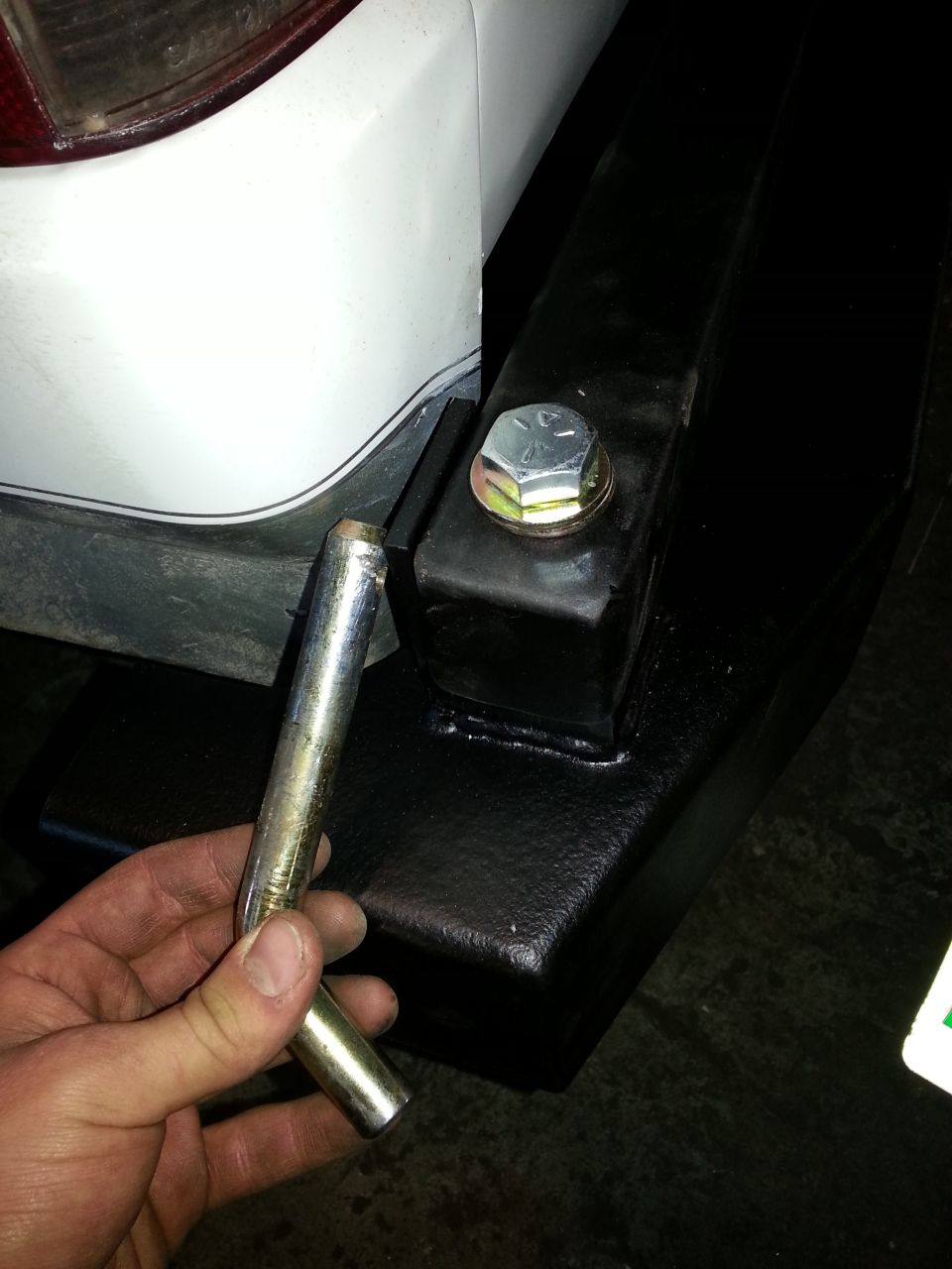 White vehicle rear bumper with black trailer hitch and a hand holding a metal pin