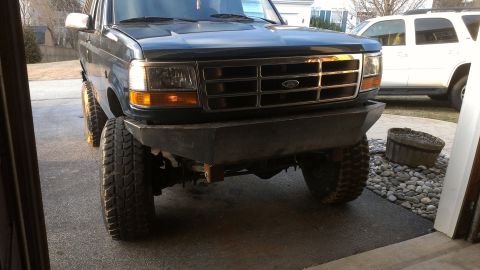 1995 Ford Bronco - A dark green Ford Bronco with an aftermarket bumper and large tires is parked in front of a white SUV.