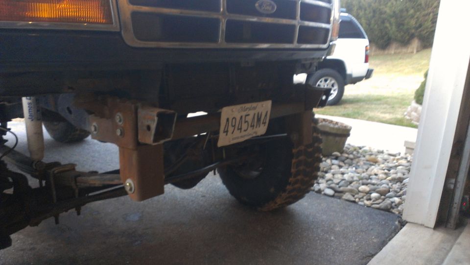 Custom bumper mount attached to the frame of a truck, with a license plate visible. Another white SUV is parked in the background