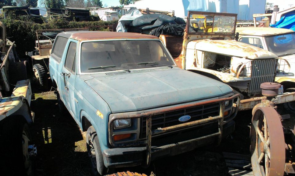 Blue Ford Bronco with a rusty roof and front brush guard, parked next to other vehicles in a junkyard