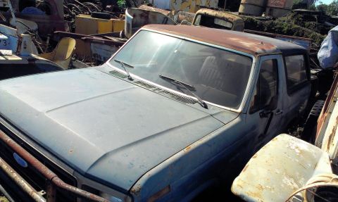 1994 Ford Bronco - This light blue Ford Bronco, likely from the 1980s, sits in a junkyard with significant rust on its roof.