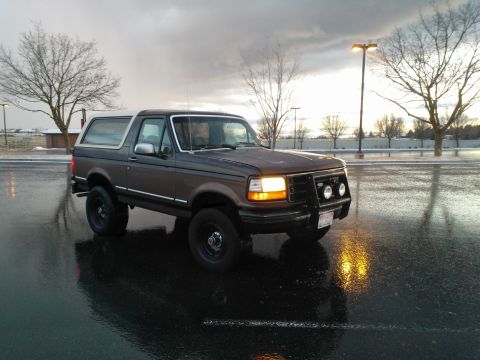 1992 Ford Bronco - A 1992 Ford Bronco sits on a wet road under a cloudy sky, reflecting the headlights.