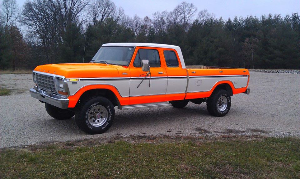 Orange and white 1970s Ford SuperCab 4x4 pickup on gravel lot, front three-quarter view