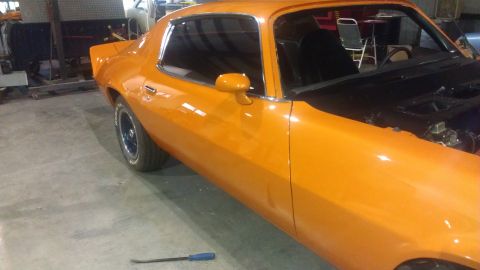 1973 Chevrolet Camaro - This orange coupe is parked inside a garage with the hood open, revealing the engine bay.