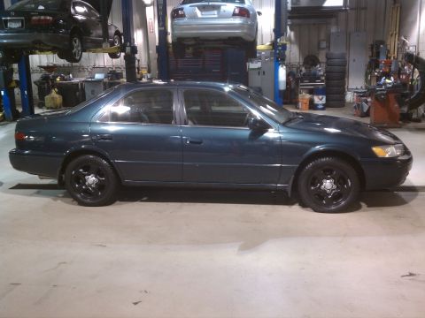 1998 Toyota Camry - A dark green sedan is parked inside a garage with another vehicle elevated above it.