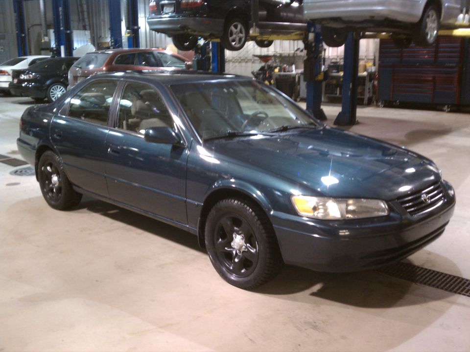 Dark teal sedan with black wheels in a garage, front 3/4 view. Another car is lifted above