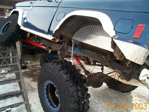 1977 Ford Bronco - A blue lifted Ford Bronco with white flares and diamond-plate rocker armor is flexed up on a ramp, exposing its underside: leaf springs, a…