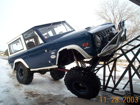 1977 Ford Bronco - Dark blue and white early Ford Bronco flexing its passenger-side front wheel up a steel ramp/flex ramp, showing coil-over shocks, radius-ar…