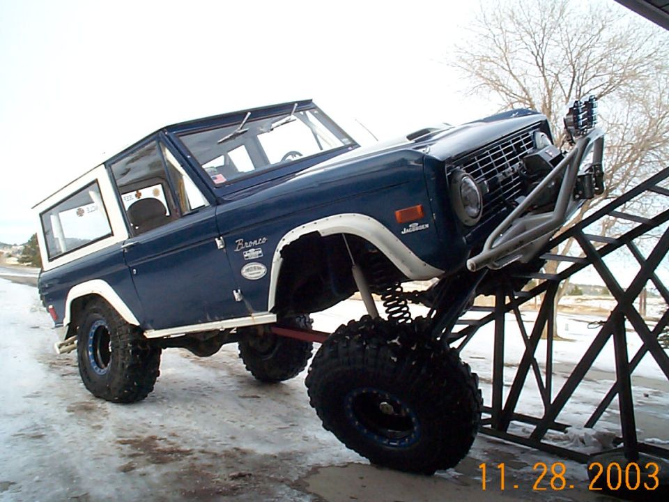 Lifted blue and white early Ford Bronco flexing front wheel on a steel ramp