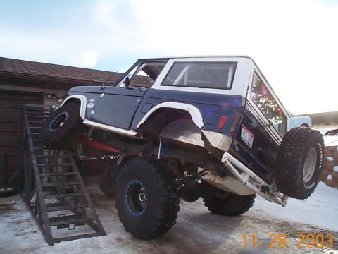 1977 Ford Bronco - Blue and white early Ford Bronco climbing a steel flex ramp with the front driver-side tire lifted, exposing the rear axle, leaf springs, d…