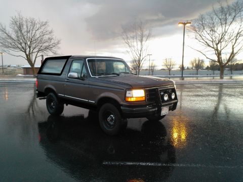 1992 Ford Bronco - A dark brown Ford Bronco is parked on a wet road under a cloudy sky.