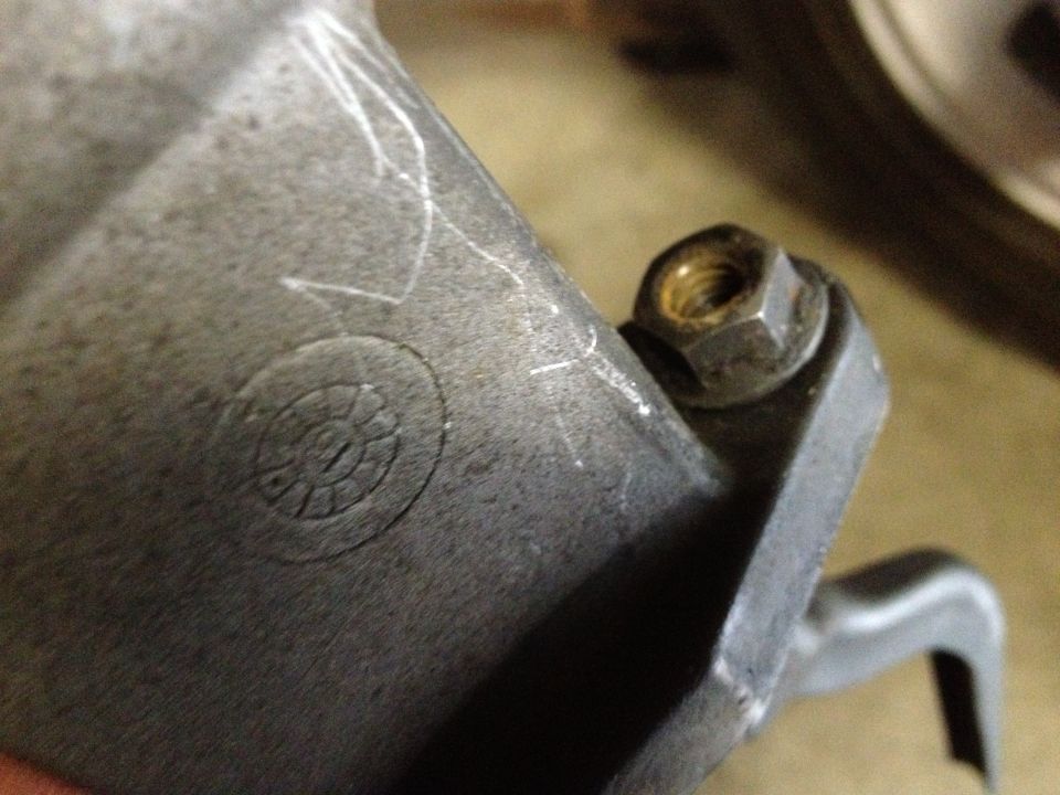 Close-up of a metal automotive part showing a bolt and nut, with some scratch marks and a circular embossed symbol