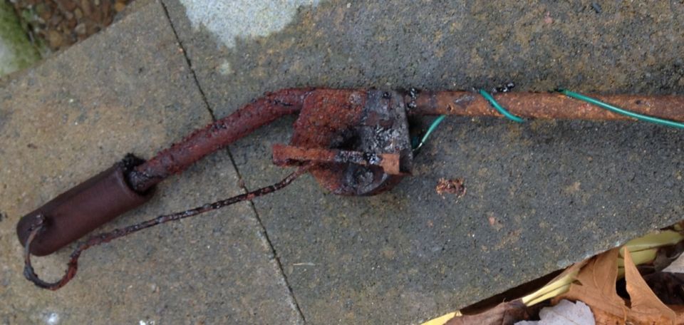 Rusted metal pipe with a cylindrical end and green wires attached, lying on concrete
