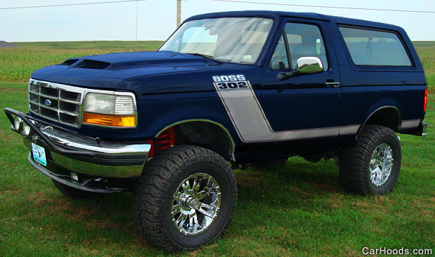 Lifted dark blue Ford Bronco with hood scoop and chrome wheels in a grassy field